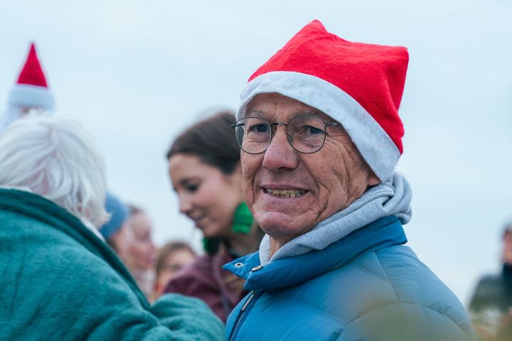 Elderly man in Santa hat and blue jacket holding a wine glass, people in background.