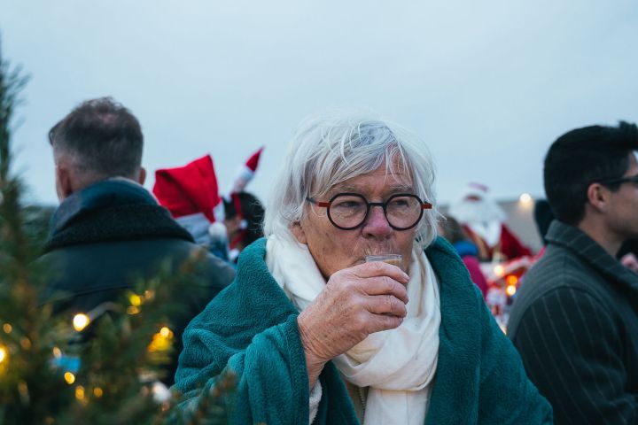 Elderly person in teal coat and glasses drinking at outdoor holiday gathering.