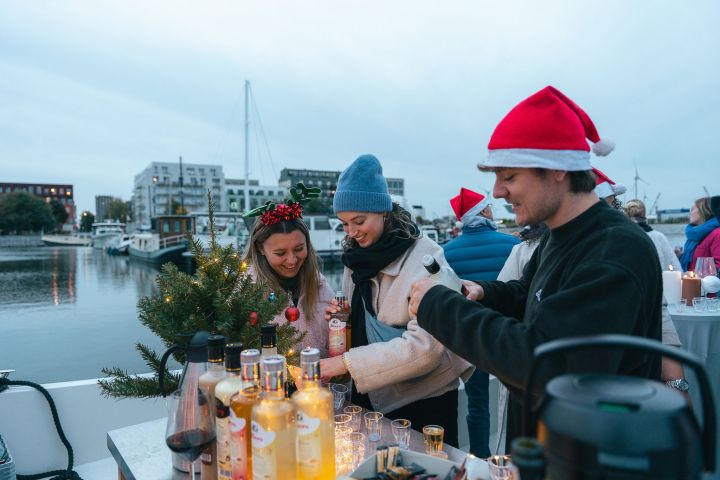 Three people in festive hats serving drinks by a marina with bottles and a small tree.