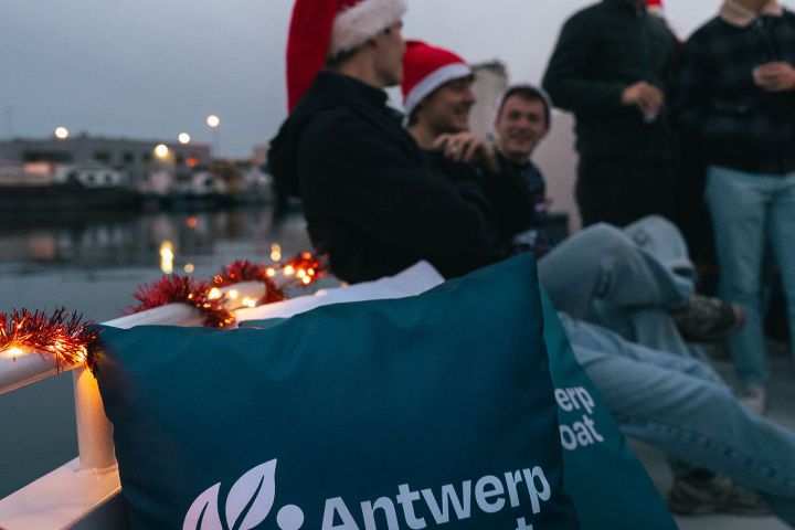 People wearing Santa hats sit on a boat with 'Antwerp by boat' pillow.