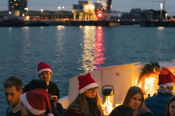 People wearing Santa hats on a boat with city lights and bridge in the background at dusk.