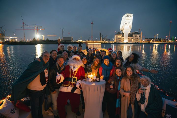 Group of people with a Santa figure on a boat by a lit building, evening setting with calm waters.