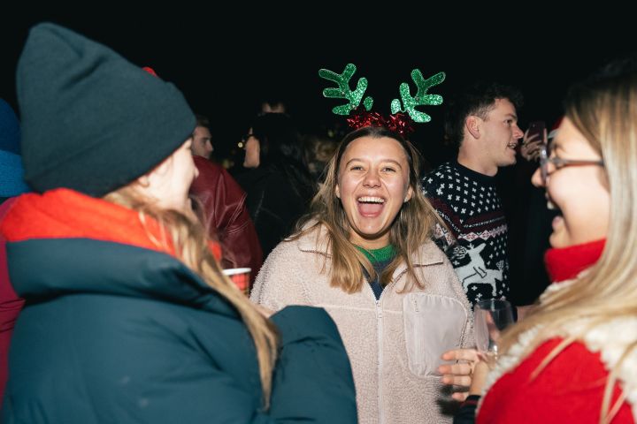 Group of people at a festive party, one wearing reindeer antlers, laughing and holding drinks.