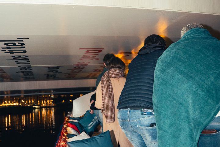 People ducking on a boat under a low bridge with colorful pillows and city lights in the background.