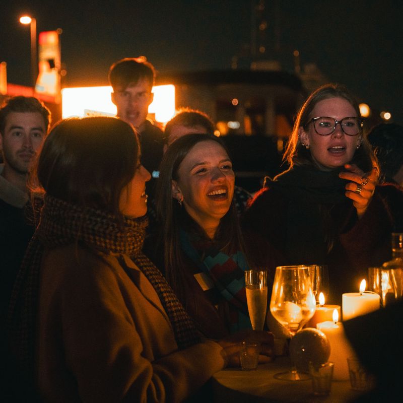 People at a night event, gathered around a table with lit candles and drinks, smiling and talking.