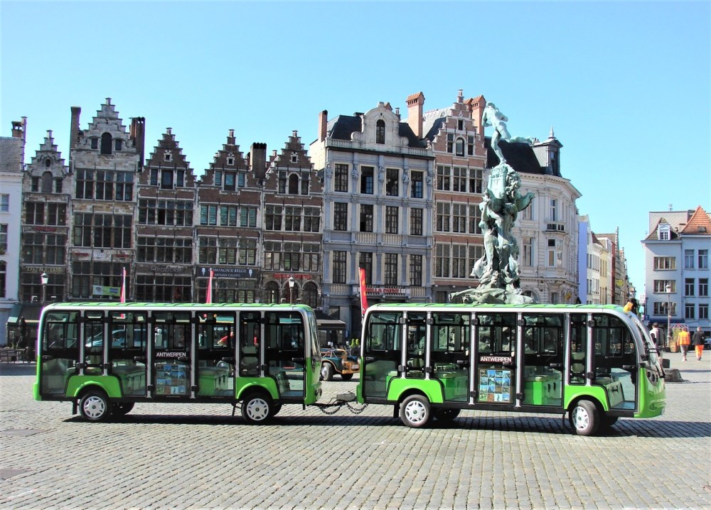 Connected green trams in a cobblestone square with historic buildings in the background.