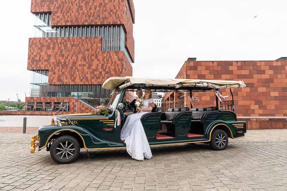 Bride and groom sit in an open vintage-style car in front of a modern brick building.