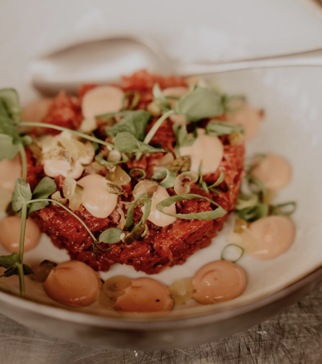 Gourmet dish with red tartare, green leaves, sauce and a spoon on a white plate.