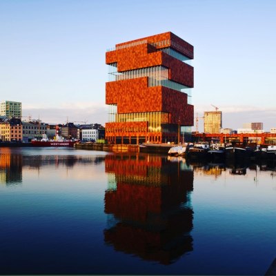 Modern stacked brick building reflecting on calm waterfront at sunset.