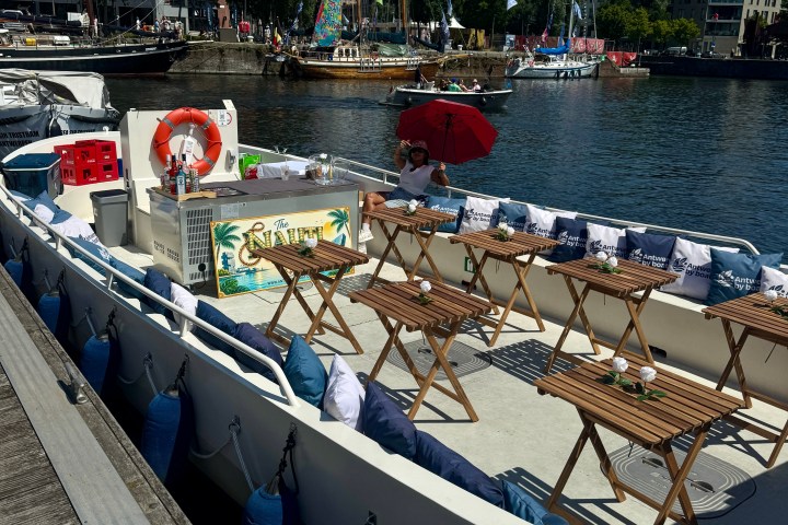 Docked boat with wooden chairs and tables, near a harbor with sailboats.